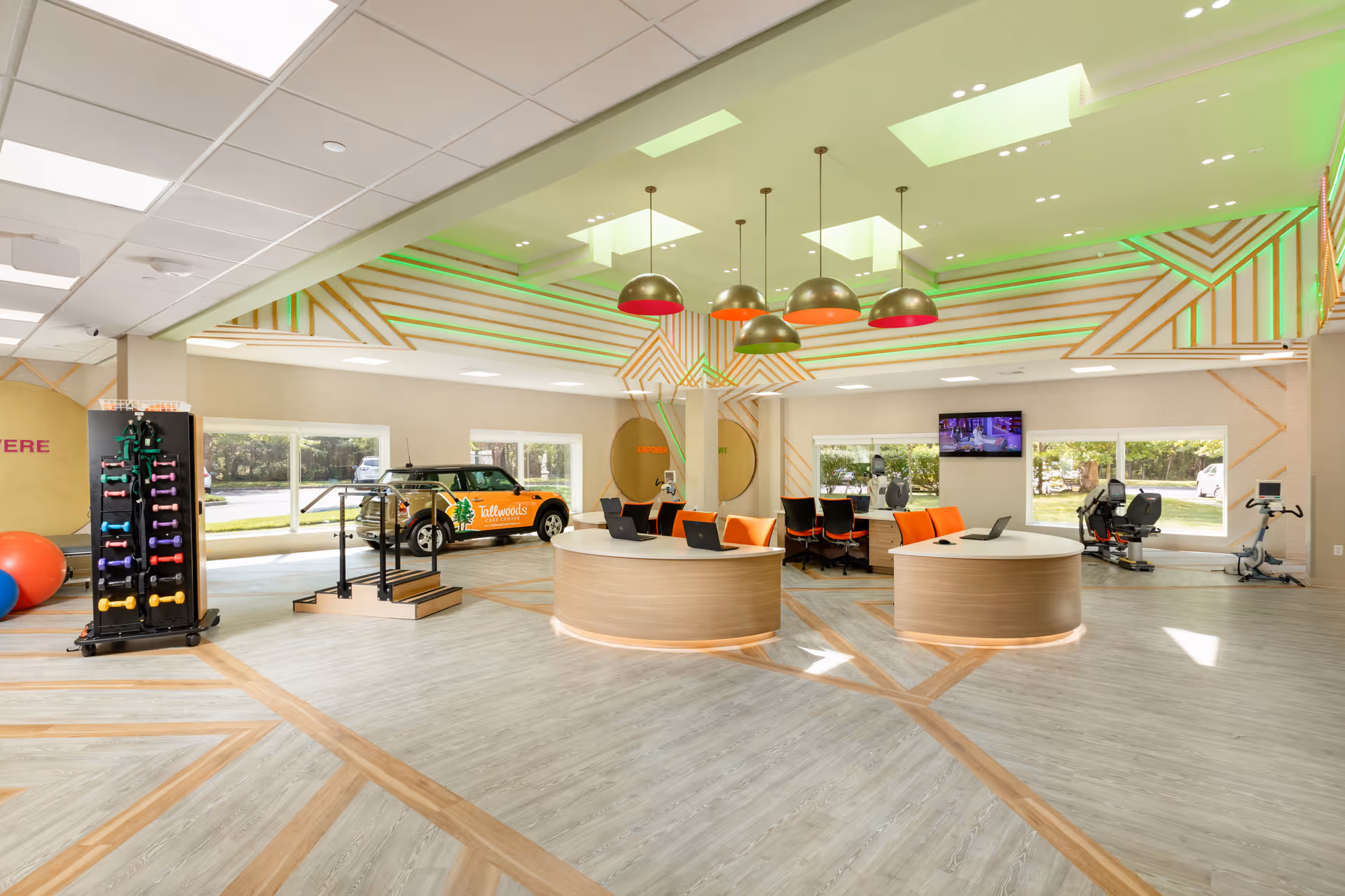 A spacious, modern rehabilitation or therapy room with light wood flooring and geometric ceiling design accented by green lighting. The room features two round reception desks with laptops and orange chairs, exercise equipment including hand weights, parallel bars, and stationary bikes. Large windows provide natural light and a view of the outdoors. A vehicle with Tallwoods branding is visible through the windows.