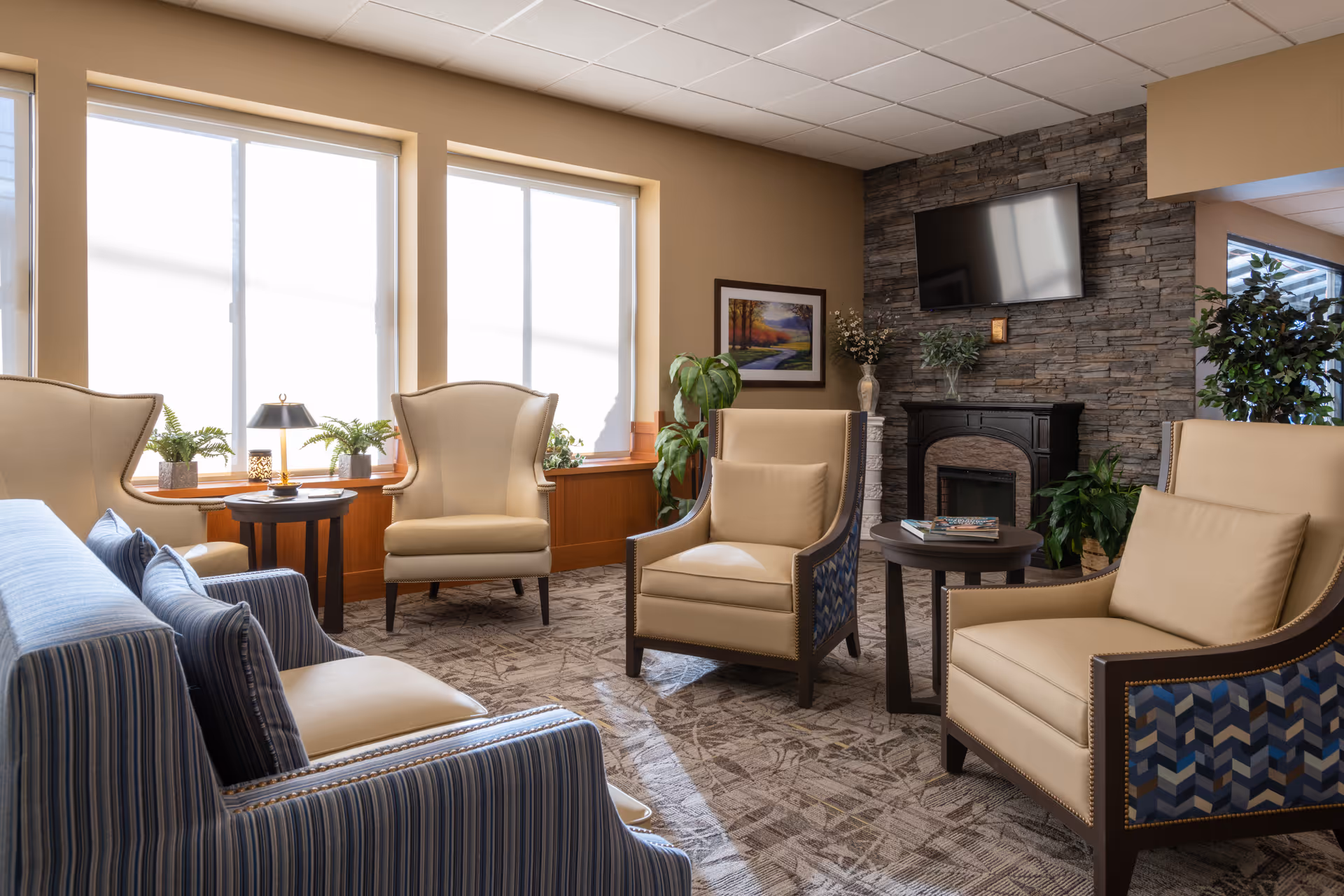 A cozy living room area in a senior living facility with beige armchairs and a striped sofa arranged around small round tables. The room features large windows with white blinds, a stone accent wall with a mounted flat-screen TV above a black fireplace, and several potted plants and framed artwork on the walls.