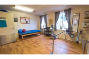 A therapy or exercise room with wooden flooring, a blue padded bench, exercise balls, a stationary bike, and large windows with curtains allowing natural light to enter.