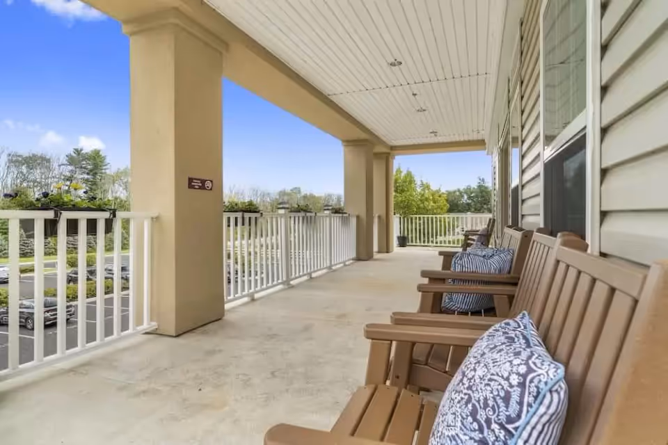 A spacious covered outdoor balcony with wooden chairs lined up along the wall, each with decorative cushions. The balcony has white railings with flower boxes attached and overlooks a parking lot and greenery under a clear blue sky.