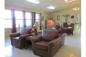 A bright living room area in a nursing home with large windows letting in natural light. The room features two brown leather armchairs with a small wooden side table and lamp between them. In the background, there is a television on a stand, a wooden piano, and a dining area with tables and chairs. The walls are decorated with mirrors and clocks.