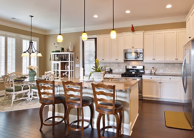 Bright and spacious kitchen with white cabinetry, granite countertops, and stainless steel appliances. A kitchen island with three wooden bar stools is in the center. Pendant lights hang above the island. In the background, there is a dining area with a glass-top table and chairs near large windows with white shutters.