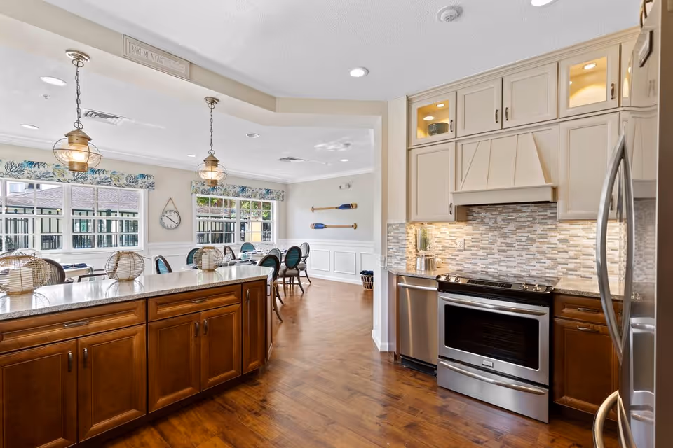 Bright and spacious kitchen and dining area with wooden floors and cabinetry. The kitchen features stainless steel appliances including a stove, dishwasher, and refrigerator. There is a large island with a granite countertop and pendant lights hanging above. The dining area has multiple tables and chairs near large windows with floral valances, and decorative oars mounted on the wall.