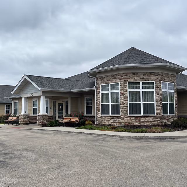 Exterior view of a single-story senior living facility building with a stone and siding facade, large windows, a covered entrance with white columns, benches outside, and a cloudy sky overhead.