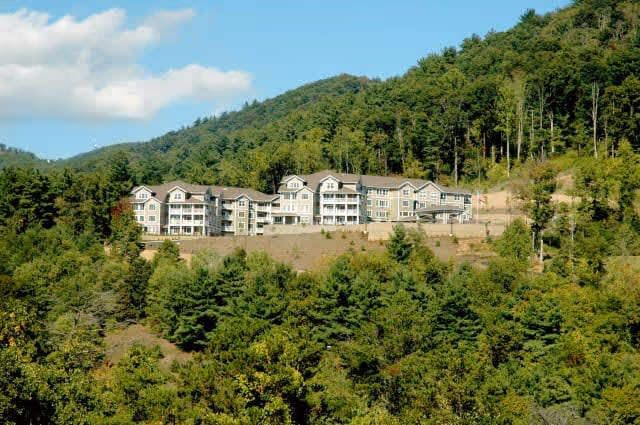 A large multi-story residential building situated on a hillside surrounded by dense green trees and forested mountains under a partly cloudy sky.