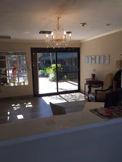 Interior view of a reception or lobby area with a chandelier hanging from the ceiling, a sliding glass door leading outside, a small round table with a chair, framed documents on the wall, and a window with decorative elements.