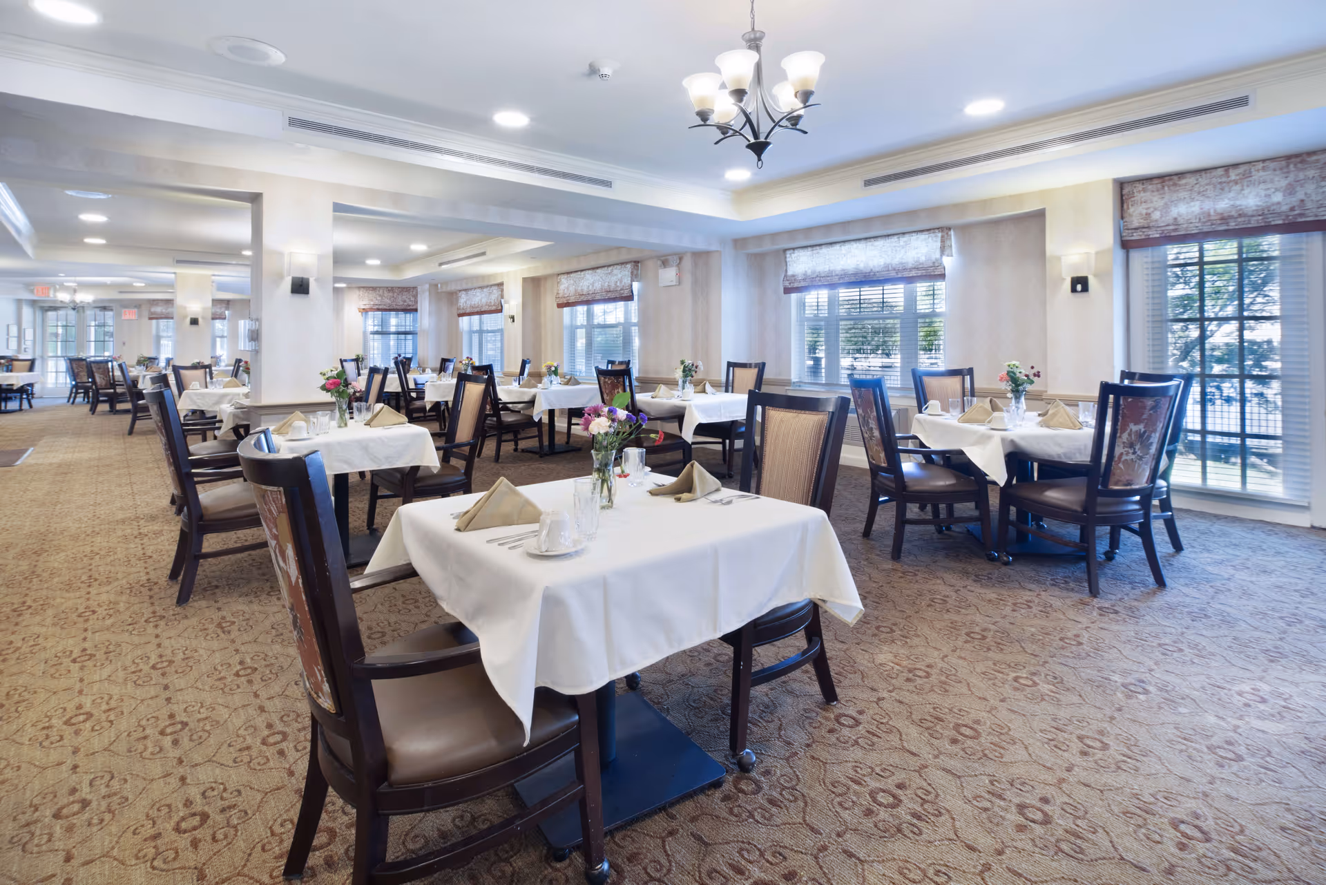 Bright, formal dining room with neatly set tables, chairs, and floral centerpieces.