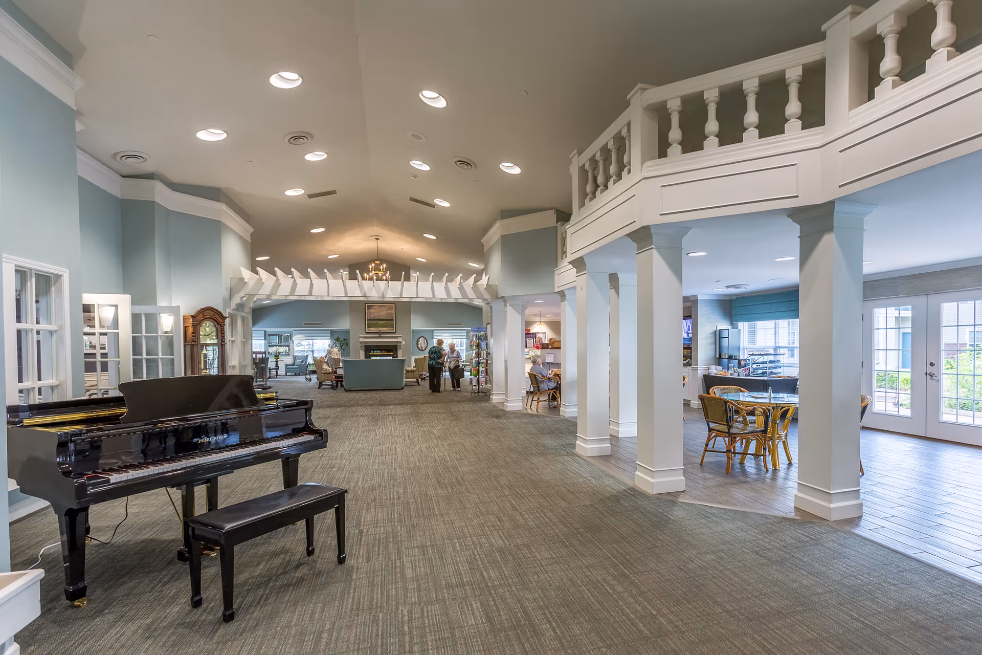 Spacious interior common area of a senior living facility featuring a grand piano, seating areas, columns, and dining tables.