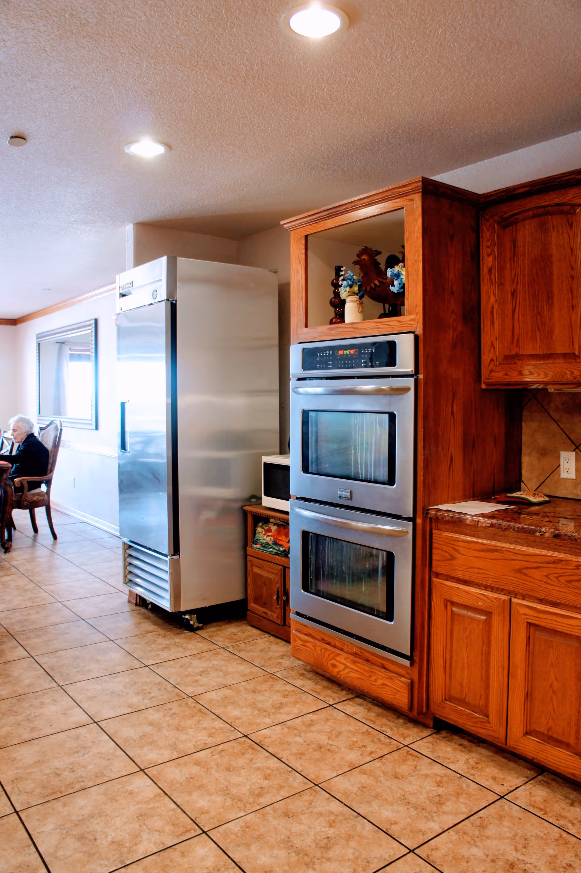 Interior view of a kitchen area with tiled floor, wooden cabinets, a double wall oven, a large stainless steel refrigerator, and a microwave on a wooden stand. In the background, an elderly person is seated at a dining table near a large mirror on the wall.