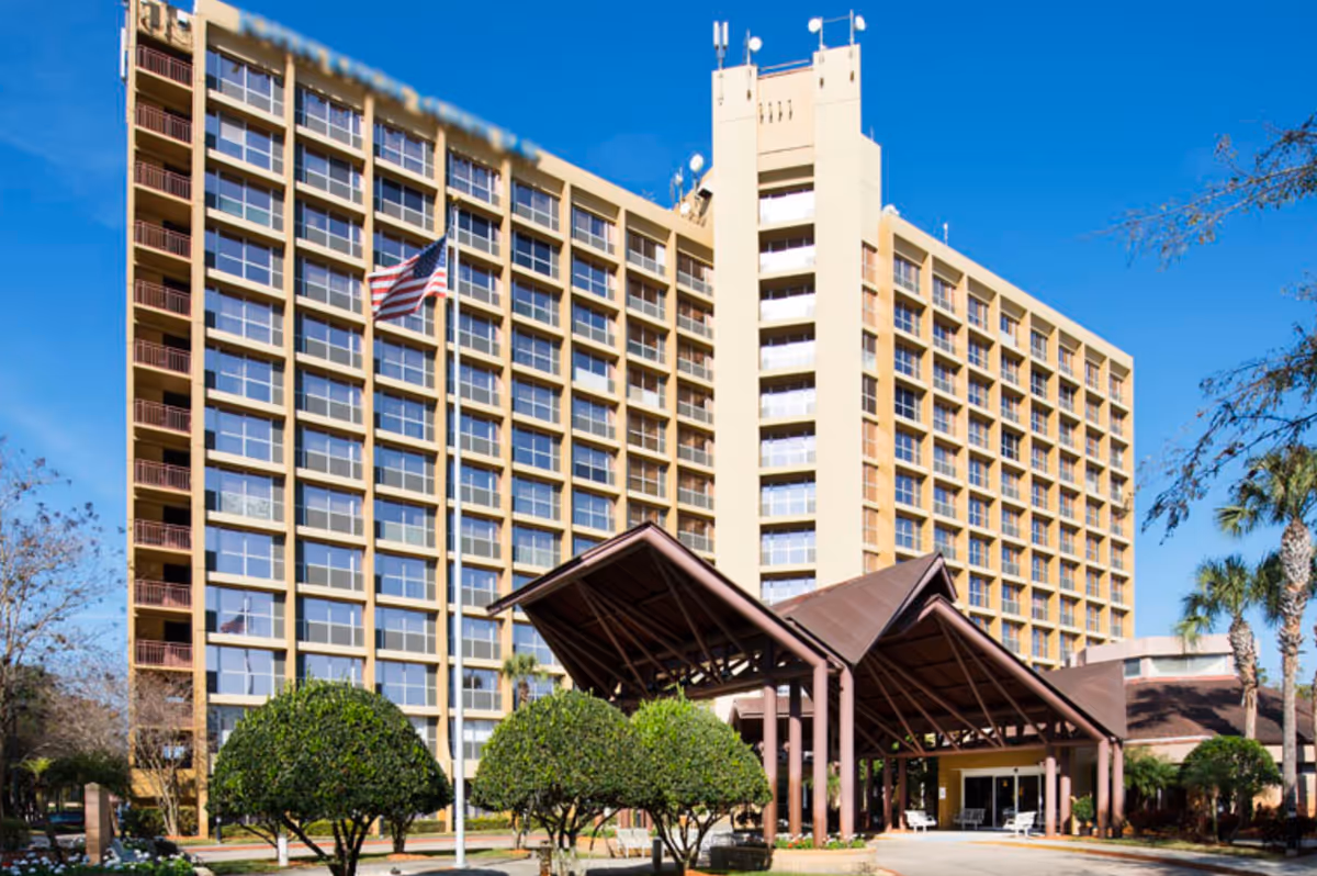 Exterior view of a multi-story senior living facility building with large windows and balconies. The entrance features a covered driveway with a brown metal roof supported by columns. There are neatly trimmed bushes and an American flag on a flagpole in front of the building, with clear blue sky in the background.