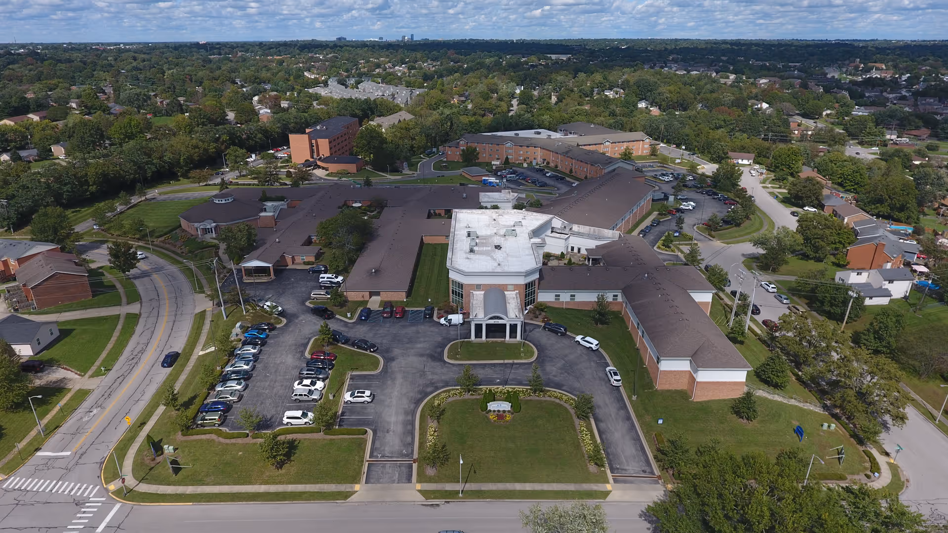 Aerial view of Sayre Christian Village, showing a large senior living facility with multiple connected buildings, parking lots with cars, surrounding greenery, and nearby residential neighborhoods under a partly cloudy sky.