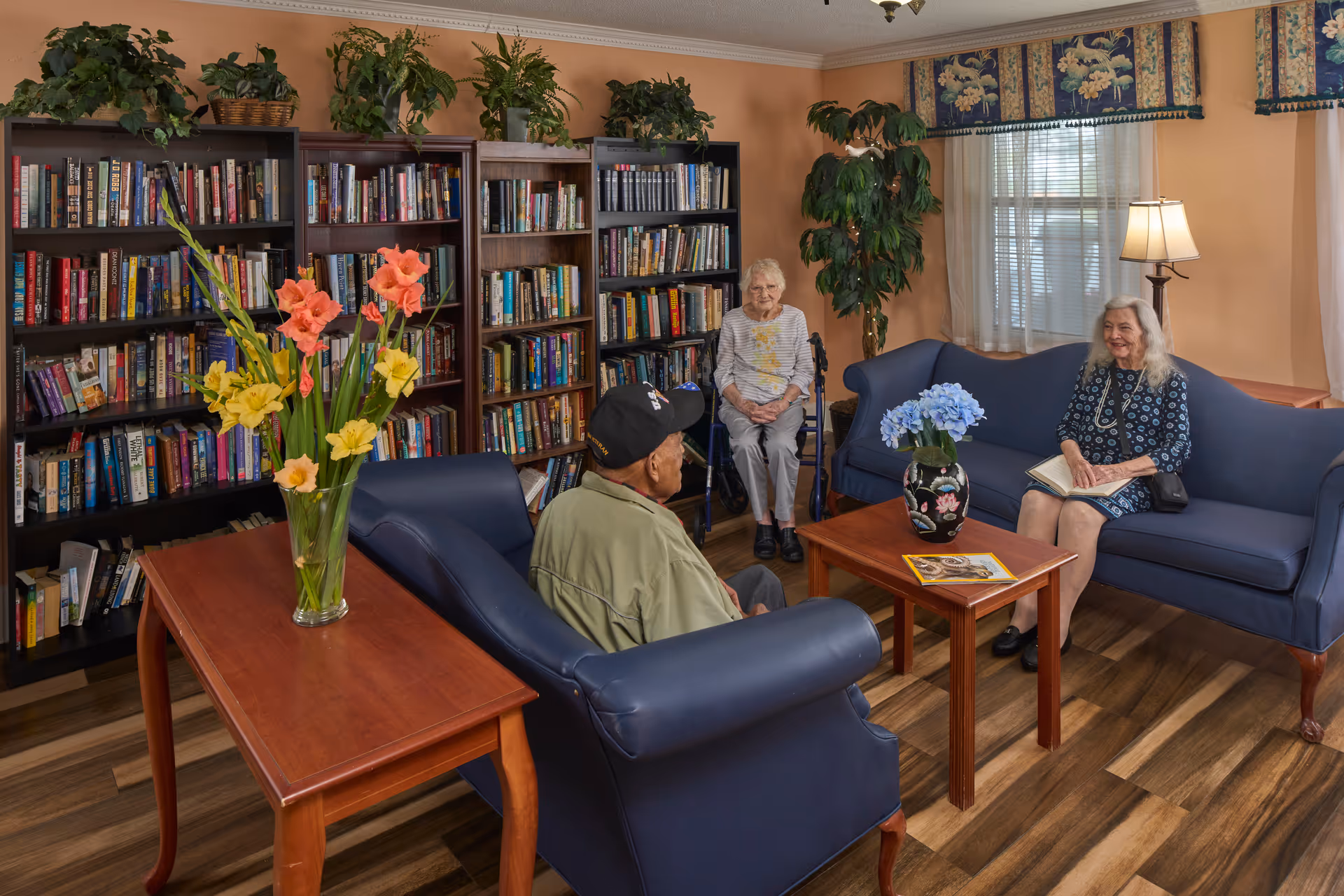 Cozy assisted living sitting room with bookshelves, blue sofas and tables with flowers, where three elderly residents are seated.