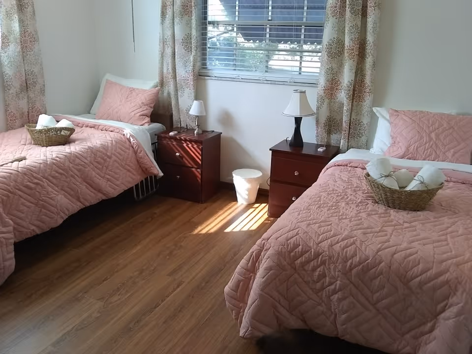Bright shared bedroom with two twin beds dressed in pink quilts, matching nightstands and lamps, a window with curtains, and baskets with towels.