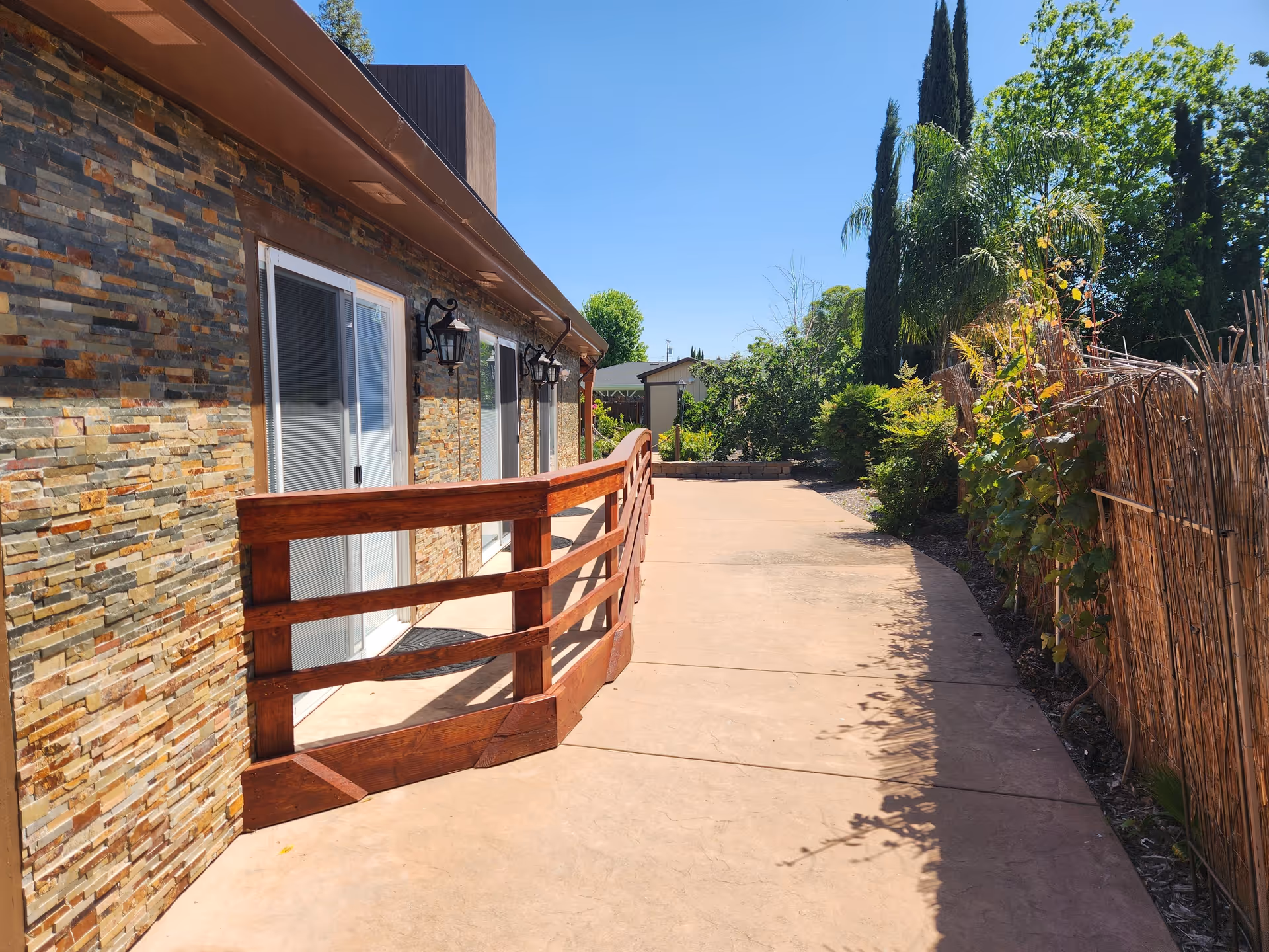 Outdoor walkway beside a building with stone exterior walls and sliding glass doors. The walkway has a wooden railing on one side and is bordered by plants and trees on the other side under a clear blue sky.