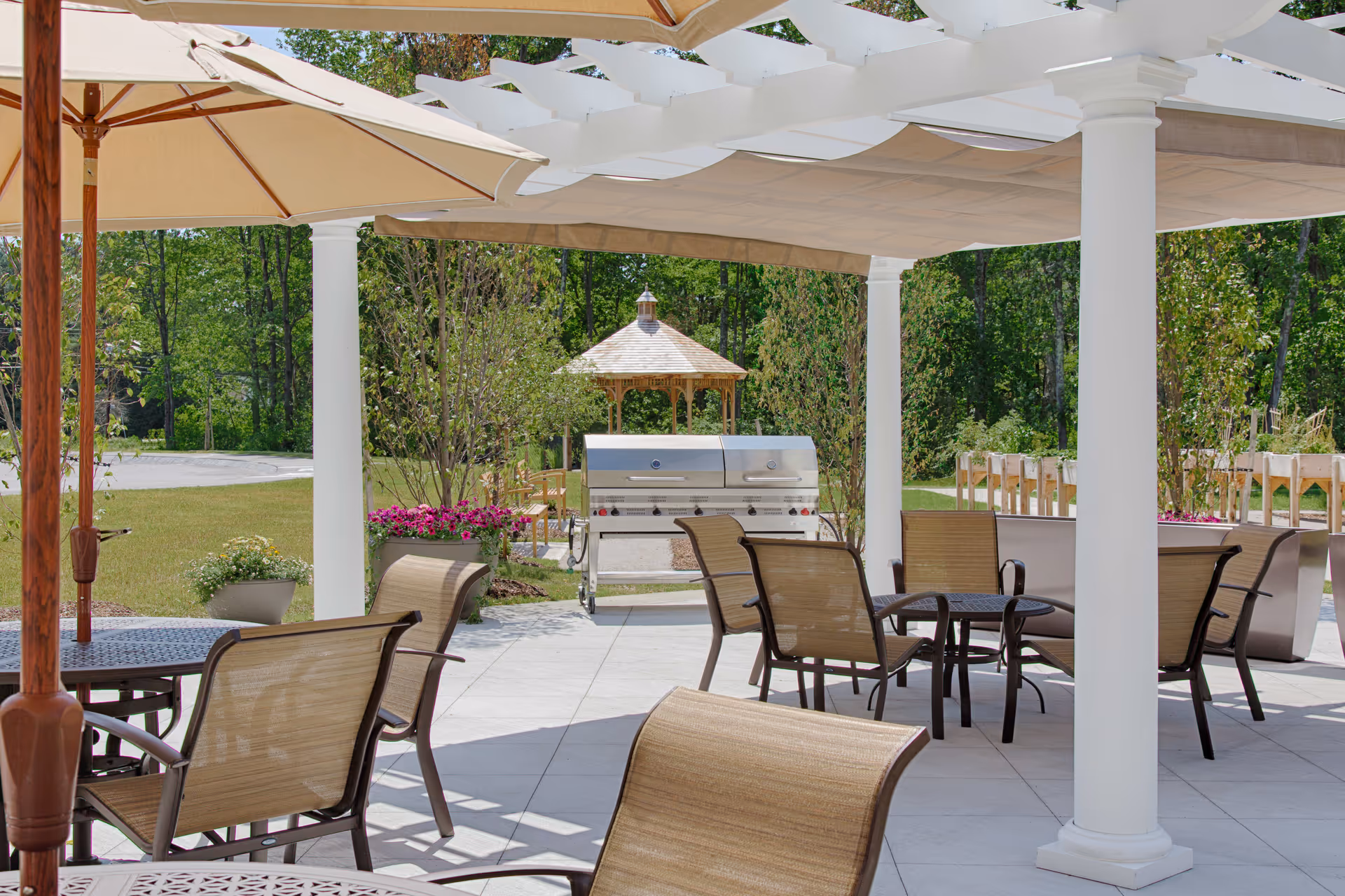 Outdoor patio area with tables and chairs under large umbrellas and a white pergola. In the background, there is a stainless steel grill and a wooden gazebo surrounded by greenery and trees.