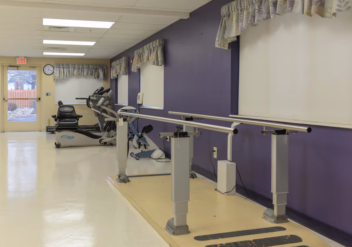 A rehabilitation room with parallel bars for walking exercises, exercise bikes, and a treadmill against a purple wall with windows covered by white blinds and floral valances. There is a door with a window and an exit sign above it at the far end of the room.