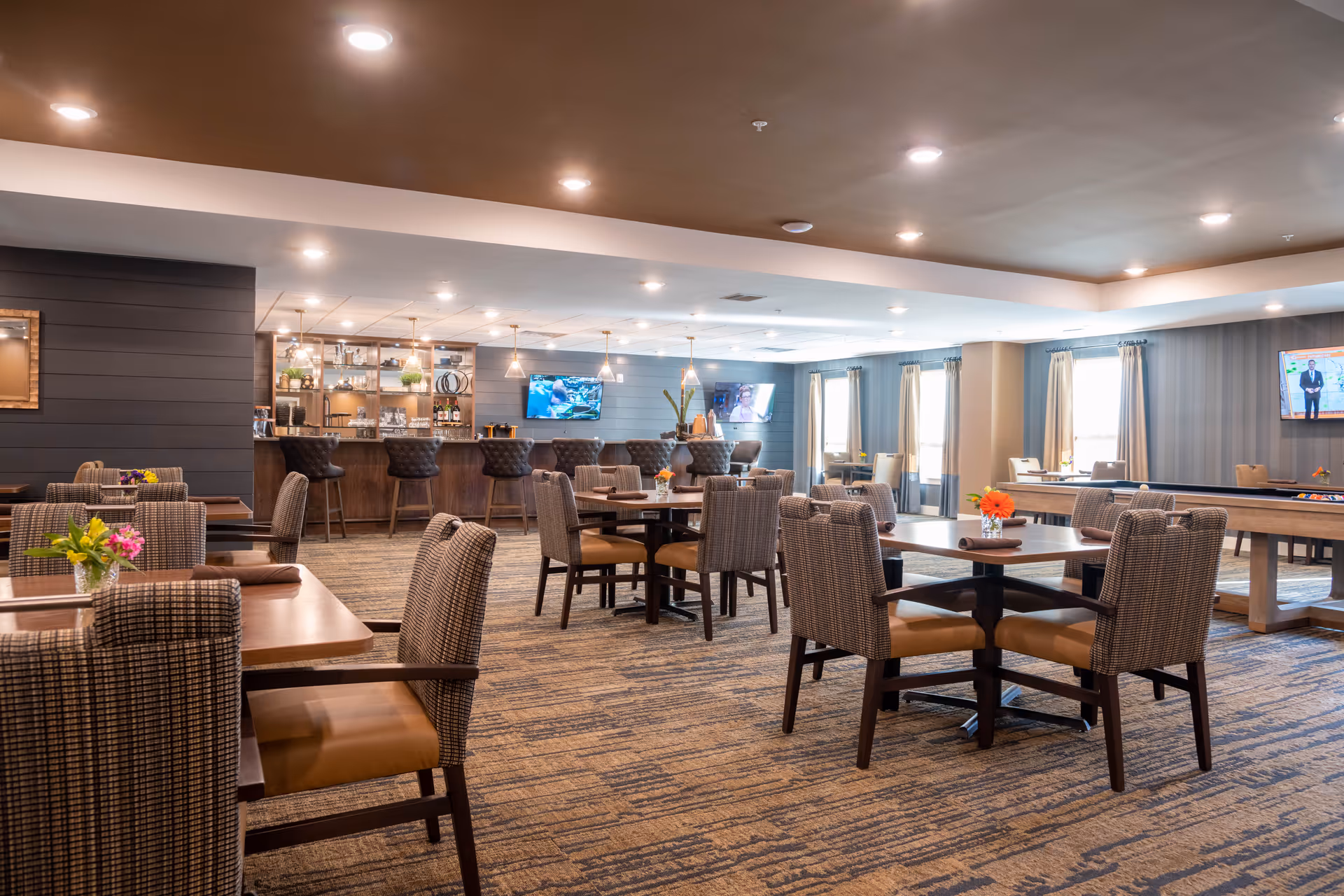 Spacious dining and lounge area with multiple tables and upholstered chairs, a bar, and wall-mounted televisions.