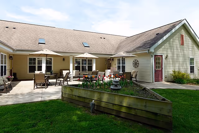 Outdoor patio area at Brookdale Battle Creek with several tables and chairs under umbrellas, surrounded by a well-maintained lawn and a raised garden bed with flowers. The building has beige siding and a brown shingled roof with skylights.