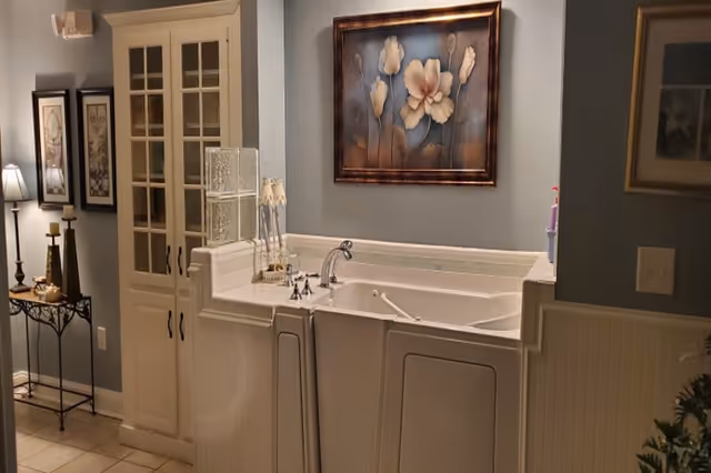 A walk-in bathtub in a bathroom with light blue walls, white cabinetry, and decorative floral artwork hanging above the tub. There are also framed pictures on the wall and a small table with candles and a lamp.
