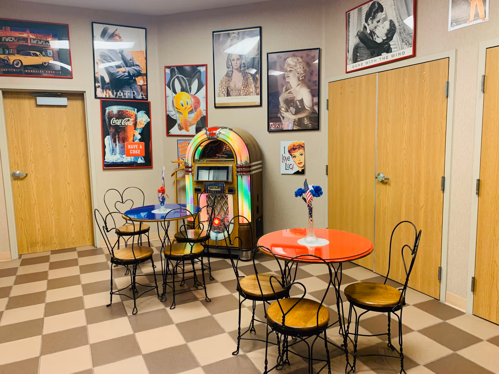 A retro-themed room with two small round tables, one red and one blue, each surrounded by black metal chairs with wooden seats. A vintage jukebox with colorful lights is against the wall. The walls are decorated with framed classic movie and cartoon posters, including images of Marilyn Monroe, Bugs Bunny, and Coca-Cola advertisements. The floor has a checkered pattern in beige and brown, and there are several wooden doors along the walls.