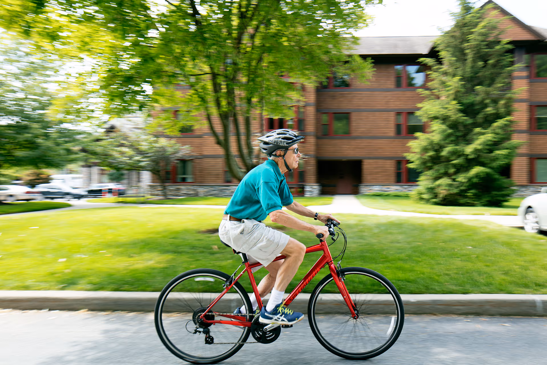 An elderly man wearing a helmet, sunglasses, a teal shirt, beige shorts, and blue sneakers is riding a red bicycle on a paved road in front of a multi-story brown building with red window frames, surrounded by green grass and trees.