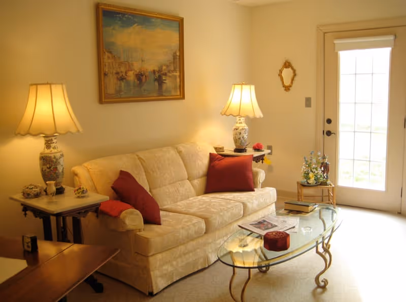 A cozy living room with a cream-colored sofa adorned with red throw pillows. Two decorative table lamps with floral bases sit on side tables flanking the sofa. A glass coffee table with a gold metal frame holds a few books and decorative items. A framed painting of boats on water hangs on the wall above the sofa. A small table with a flower arrangement is near a glass door letting in natural light.