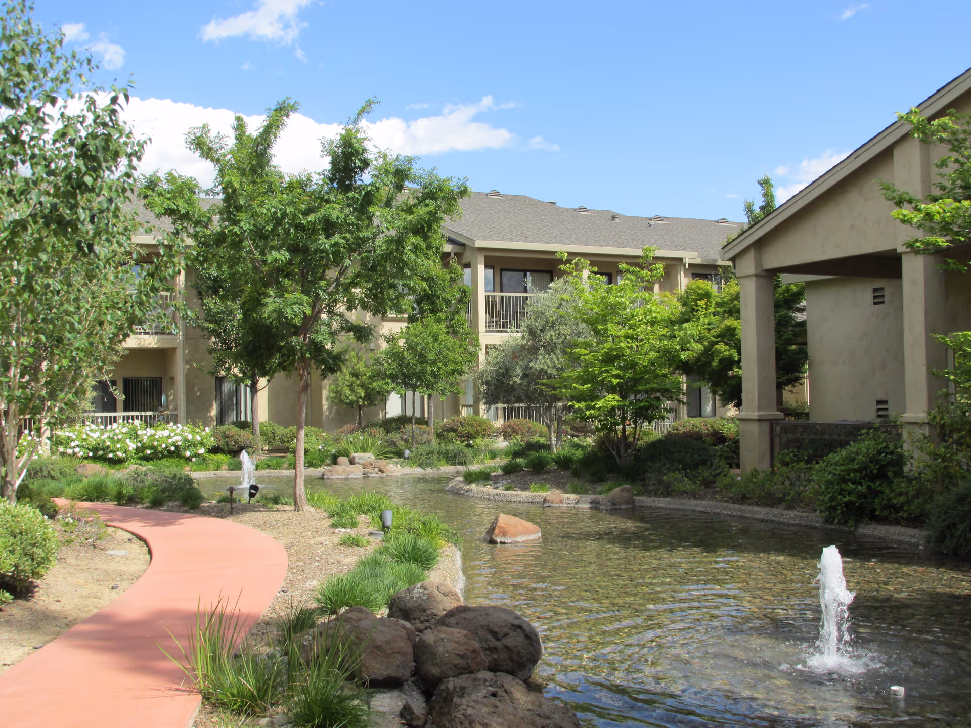 Outdoor view of a senior living facility with a winding red walking path next to a pond with small fountains. The area is landscaped with green trees, bushes, and rocks, and beige two-story buildings are visible in the background under a blue sky with some clouds.
