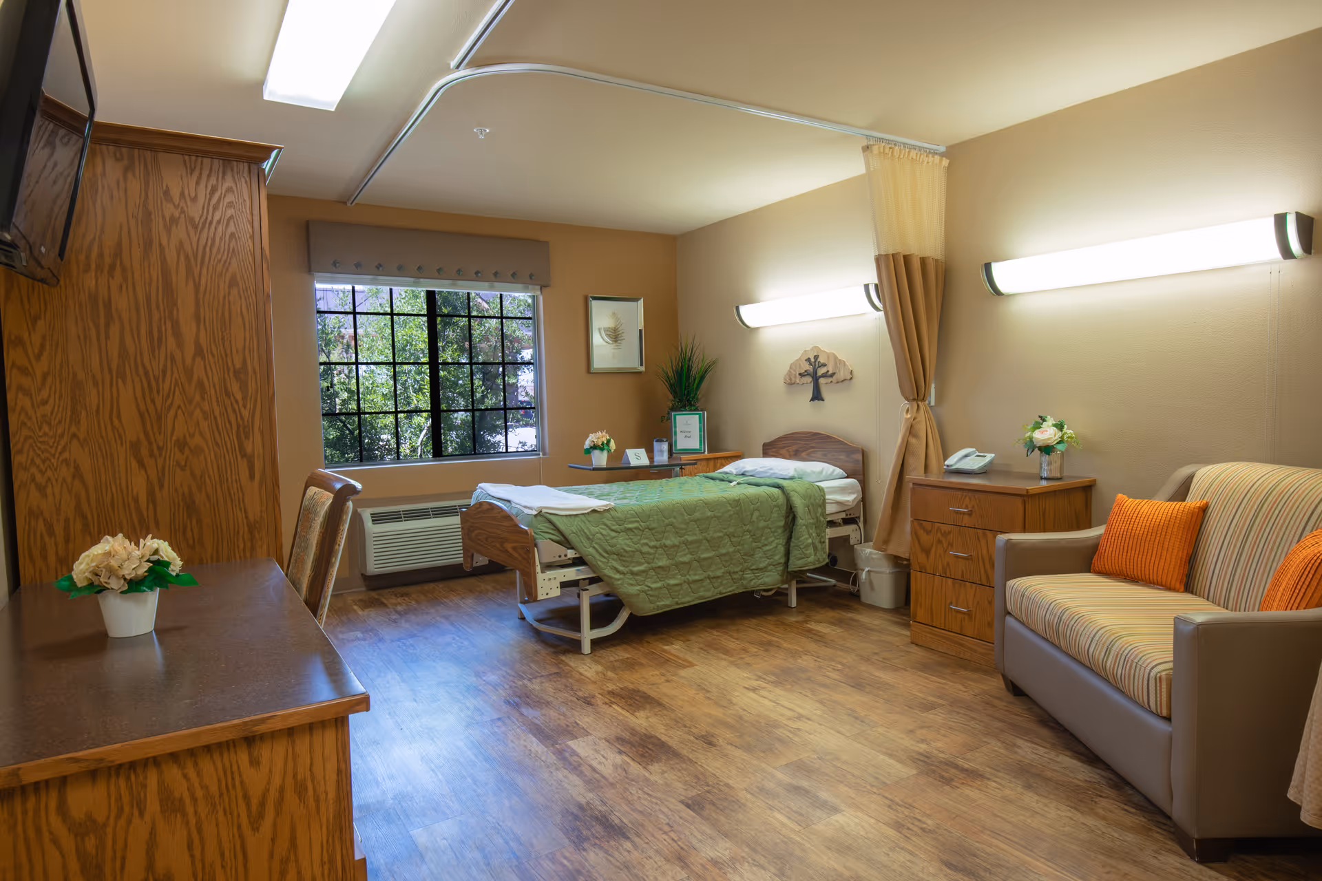 A well-lit senior living facility room with a single bed covered in a green quilt, a wooden desk with a chair, a striped sofa with orange pillows, a wooden cabinet, and a window showing greenery outside. The room has wood flooring and beige walls with wall-mounted lights.
