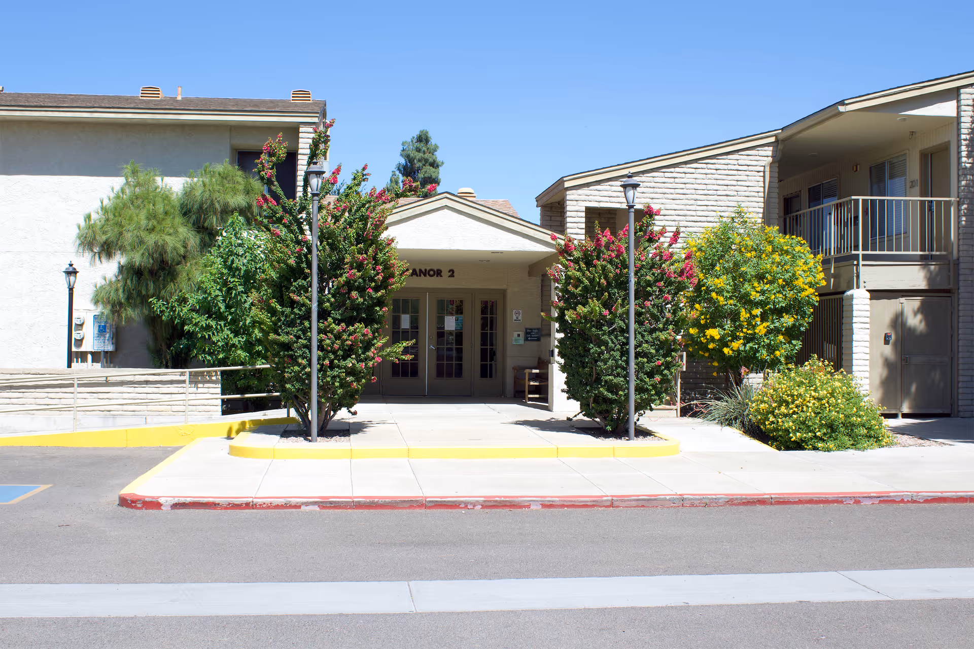 Exterior view of a senior living facility entrance labeled 'Manor 2' with two flowering trees and bushes on either side of the walkway, a clear blue sky overhead, and a two-story building with balconies on the right.