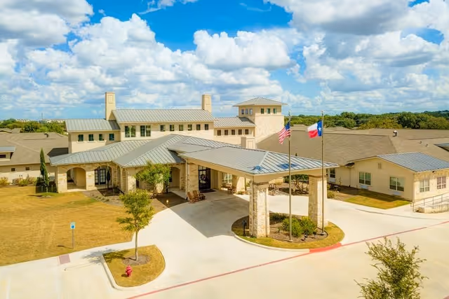 Exterior view of The Auberge at Cedar Park senior living facility showing a large building with a covered entrance, two flagpoles with American and Texas flags, surrounded by landscaped grounds under a partly cloudy sky.