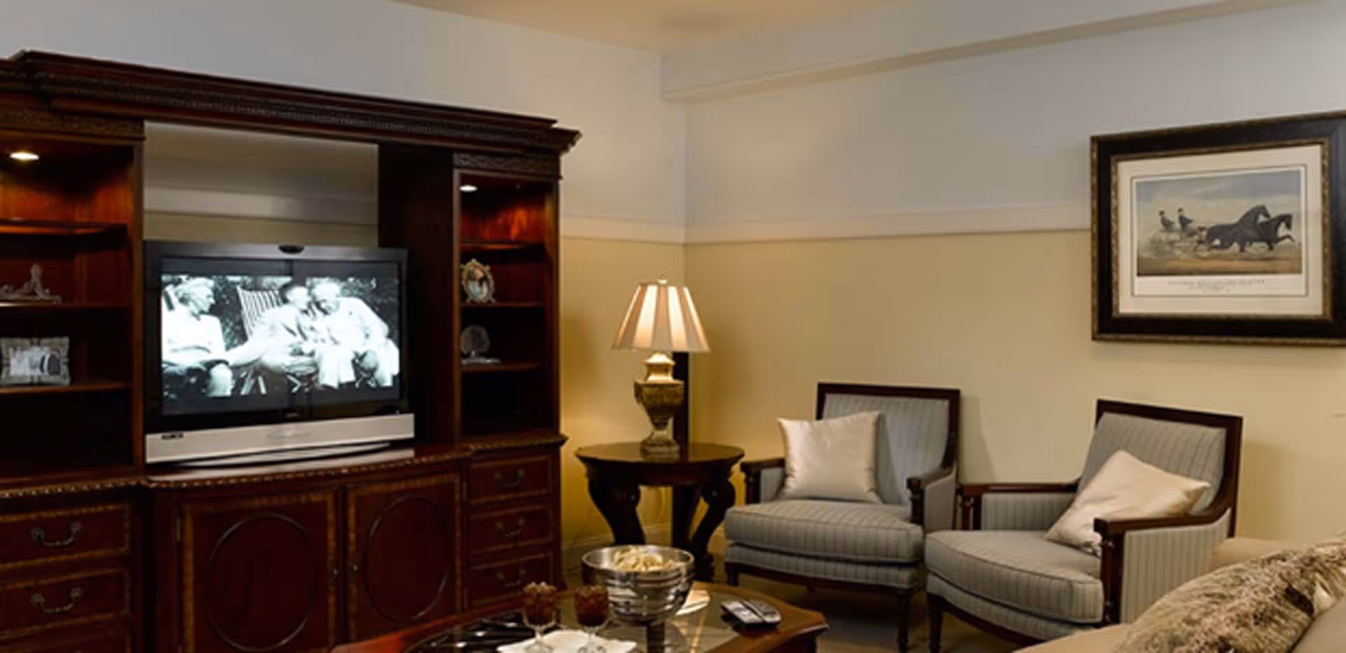 Cozy living room with a wooden entertainment center and TV, two upholstered armchairs, a side table with lamp, and framed wall art.