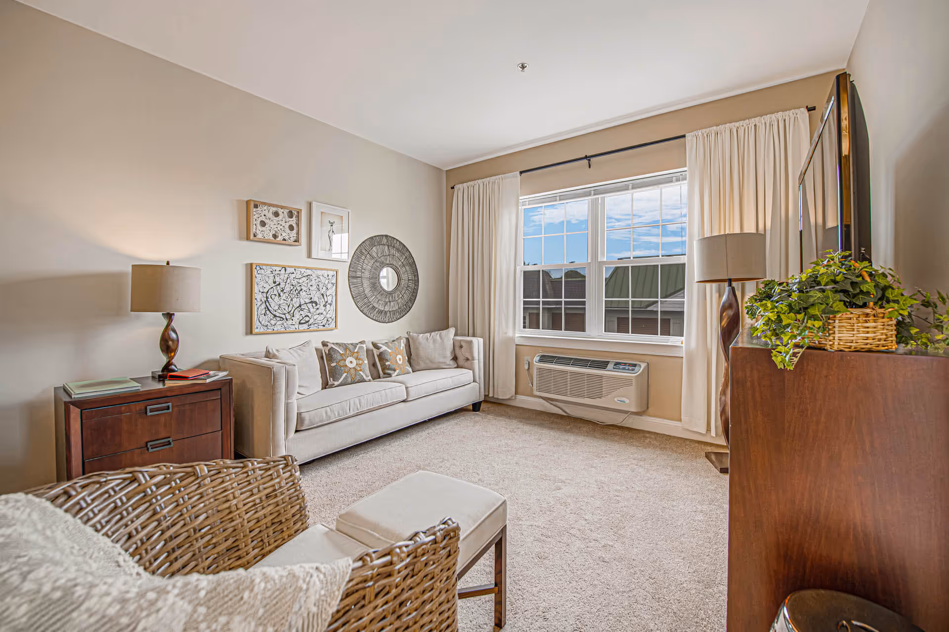A bright living room with a beige sofa adorned with decorative pillows, a wicker chair with a cushion, a wooden side table with a lamp, and a large window with cream-colored curtains letting in natural light. A wall-mounted air conditioning unit is below the window, and a wooden cabinet with a lamp and a green plant is on the right side. The walls are decorated with framed artwork and a round mirror.