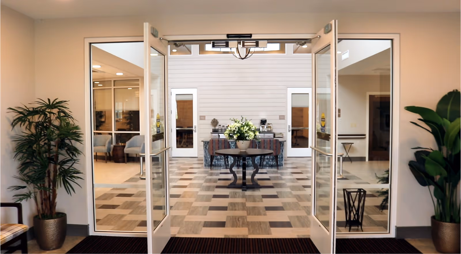 Open double glass doors leading into a bright interior lobby with a central table topped with flowers, potted plants, and seating areas.