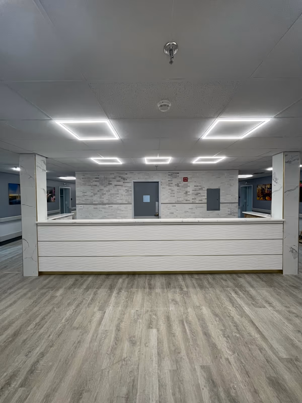 Interior view of a nursing and rehabilitation center hallway with a modern reception desk in the center. The floor has light wood-style vinyl flooring, and the walls are decorated with light-colored tiles and marble-patterned columns. The ceiling has square LED light fixtures, and there are doors and framed pictures along the hallway.