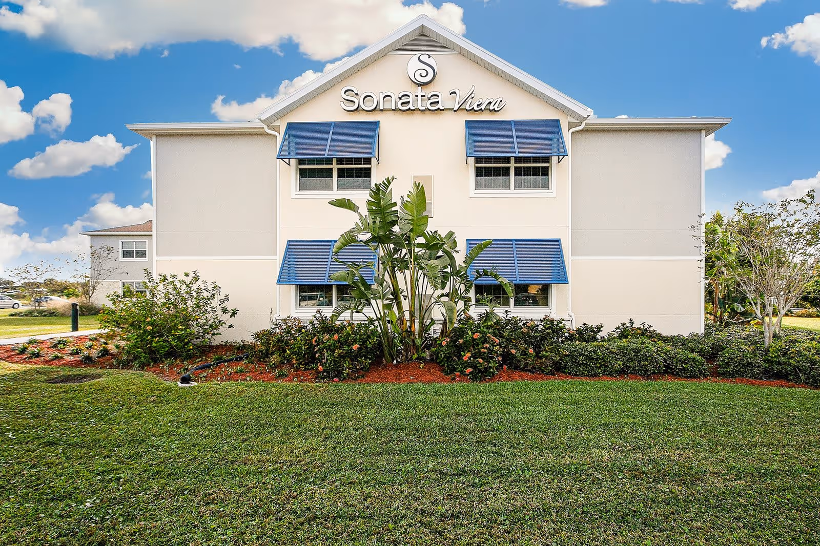 Exterior view of a two-story building with beige walls and blue window awnings, surrounded by green grass, bushes, and small trees under a partly cloudy sky. The building has a sign that reads 'Sonata Viera' at the top.