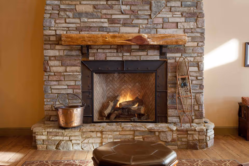 Stone fireplace with a wooden mantel and a fire burning inside. A copper bucket is placed on the left side of the hearth, and a vintage snowshoe leans against the right side. Part of a brown leather ottoman and a patterned rug are visible in front of the fireplace.