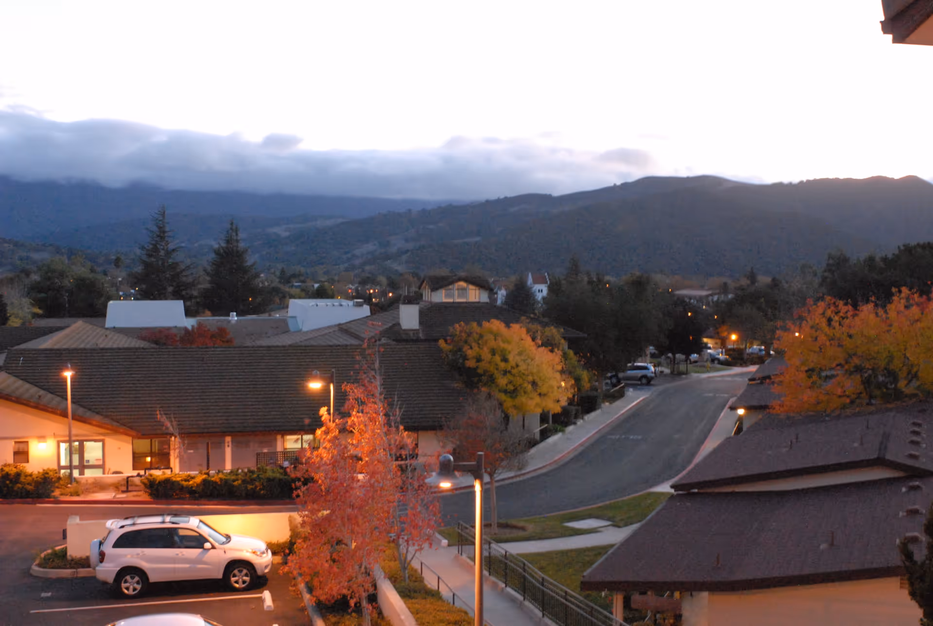 View of a senior living facility parking lot and buildings at dusk with street lamps on, surrounded by trees with autumn foliage and hills in the background under a cloudy sky.