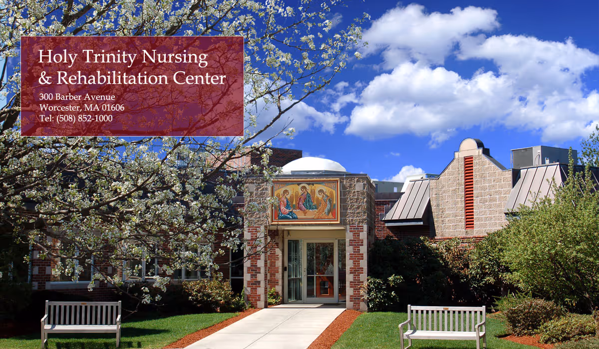 Front entrance of Holy Trinity Nursing & Rehabilitation Center with a walkway, benches, blossoming tree and blue sky.