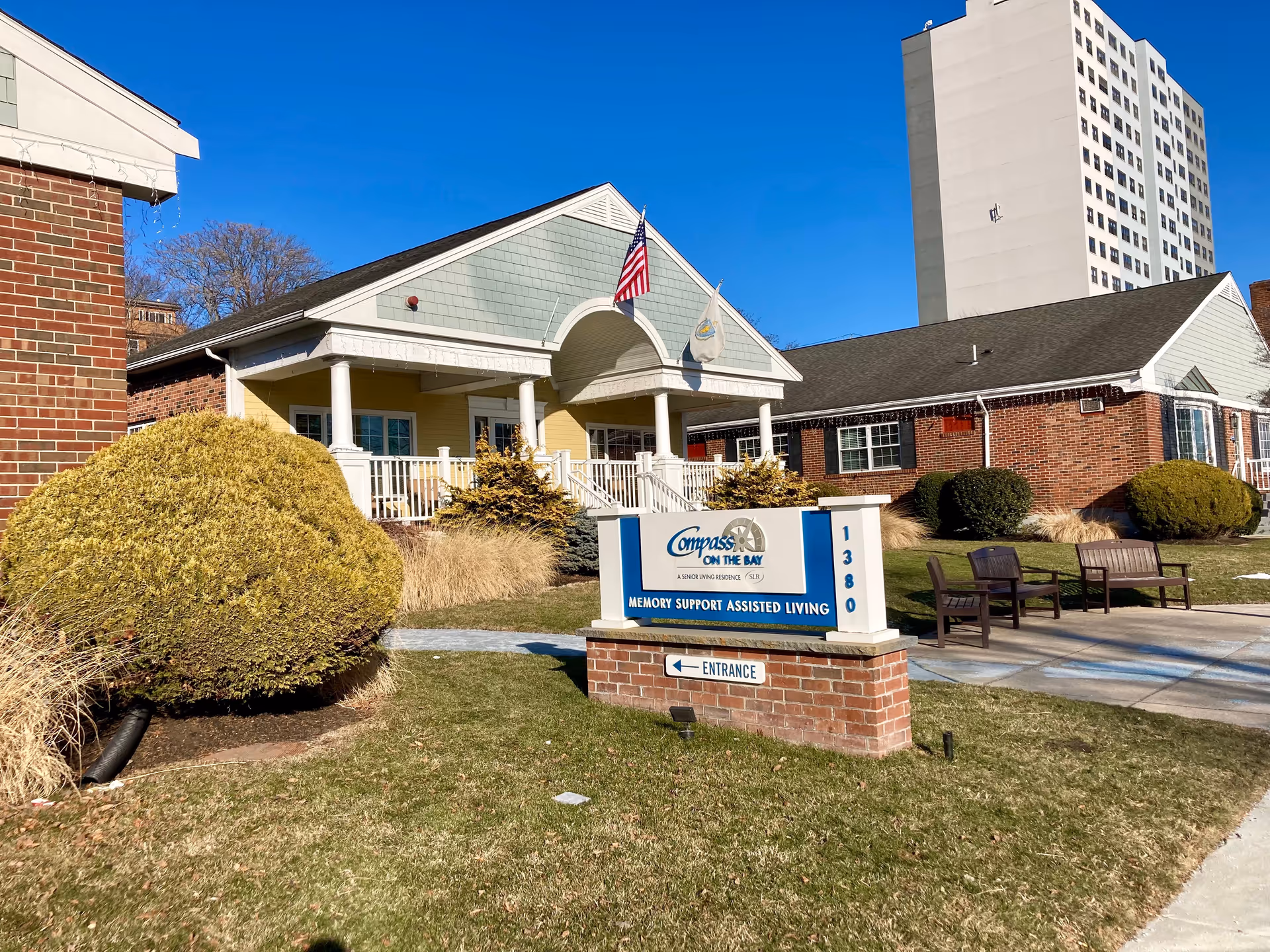 Exterior view of Compass on the Bay Memory Support Assisted Living facility showing a single-story building with a porch, two flags, bushes, and a sign indicating the entrance. There are benches on the sidewalk and a tall building in the background under a clear blue sky.
