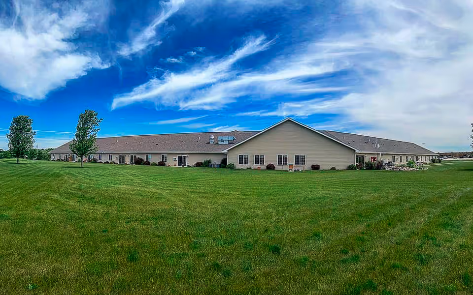 Wide exterior view of a single-story senior living facility building with beige siding and a brown roof, surrounded by a large green lawn and a few trees under a partly cloudy blue sky.