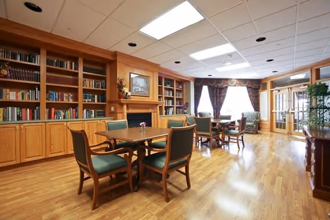 A bright and spacious common area with wooden flooring, several tables and chairs, built-in wooden bookshelves filled with books, a fireplace with a framed painting above it, large windows with dark curtains, and a glass door leading outside.