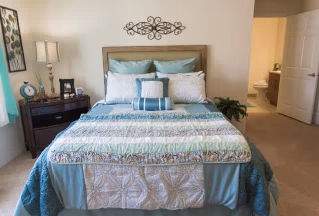 A neatly made bed with multiple pillows and a patterned quilt in shades of blue and beige, positioned against a beige wall with a decorative metal wall art above the headboard. To the left of the bed is a dark wooden nightstand with a lamp, clock, and other small decor items. An open doorway on the right side reveals a bathroom with a toilet and vanity.