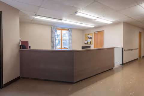 Interior view of a nursing facility reception or nurse station area with a brown counter, beige walls, a window with floral curtains, fluorescent ceiling lights, and a hallway with handrails.