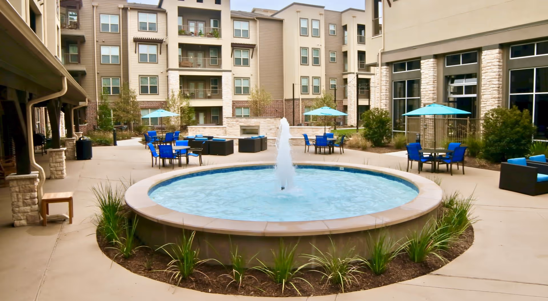 Outdoor courtyard area with a round water fountain in the center, surrounded by patio tables with blue chairs and turquoise umbrellas. The courtyard is enclosed by multi-story residential buildings with balconies and large windows. There are some plants and shrubs around the fountain and along the edges of the courtyard.