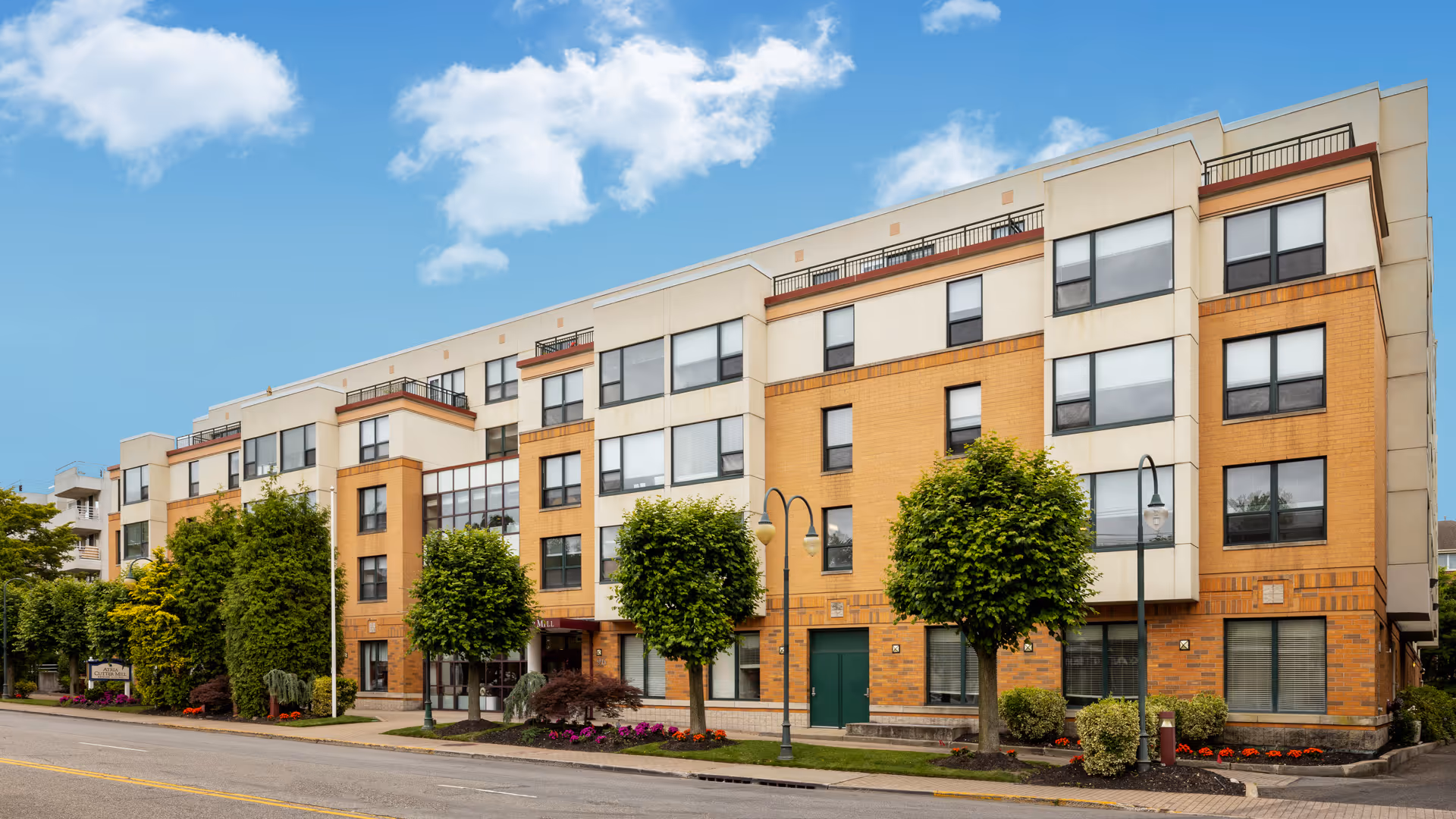 Facade of a four-story residential building with brick and light-colored panels, street trees, and landscaped flower beds under a blue sky.