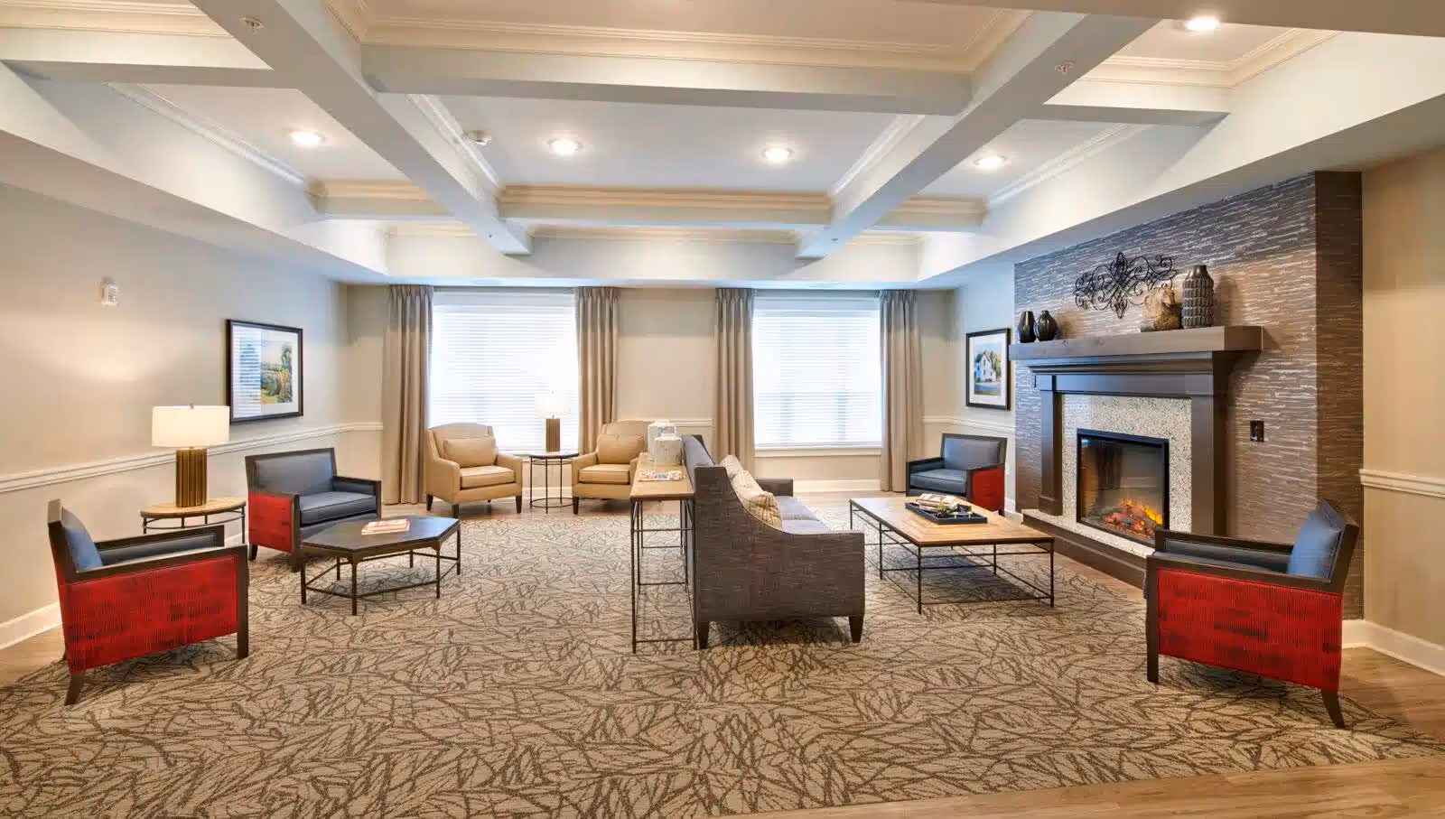A spacious and well-lit living room area with a patterned carpet, multiple seating options including armchairs with red sides, beige chairs, and a gray sofa. There is a modern fireplace with decorative items on the mantel and framed artwork on the walls. Large windows with beige curtains allow natural light to fill the room.