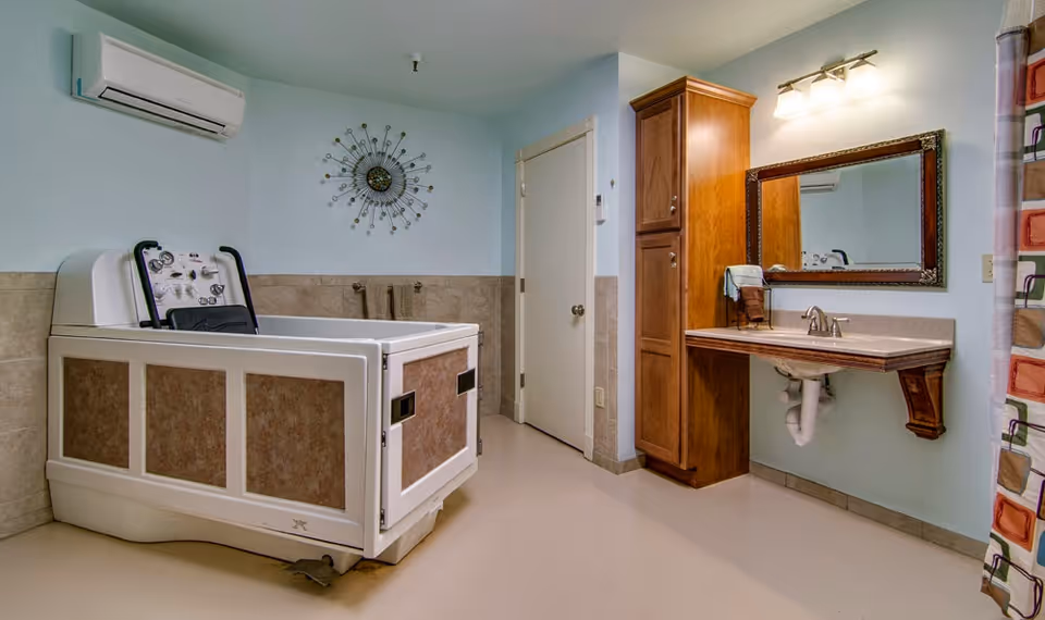 A spacious bathroom featuring a large accessible bathtub with control panel, a wall-mounted air conditioner, a decorative wall clock, a wooden cabinet, a sink with a mirror above it, and a colorful shower curtain on the right side.