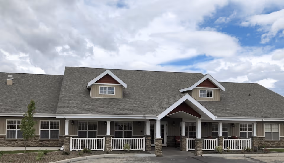 Front exterior view of a single-story assisted living facility building with a gray roof, beige siding, stone accents on the lower walls, white railings on the porch, and a partly cloudy sky in the background.