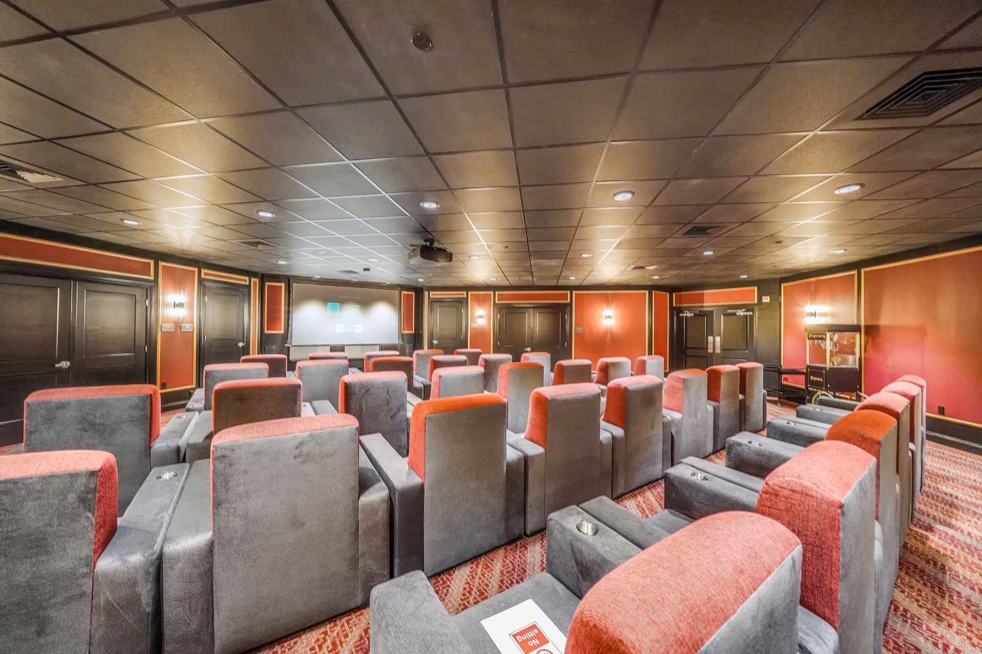 A private theater room with rows of red and gray upholstered reclining seats facing a projection screen and ceiling-mounted projector.