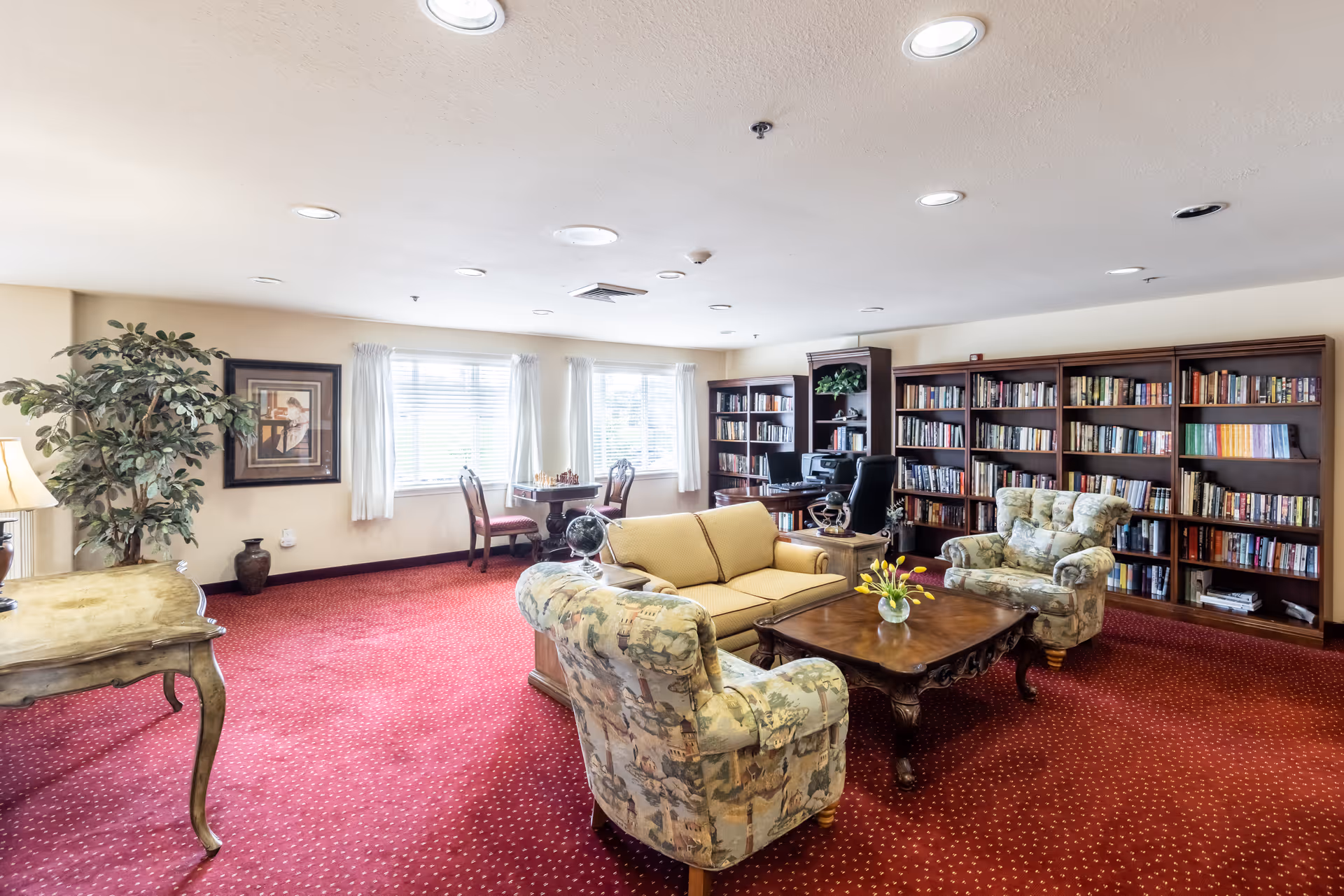 A bright senior living lounge/library with bookshelves, upholstered chairs and a sofa around a coffee table on red carpet.